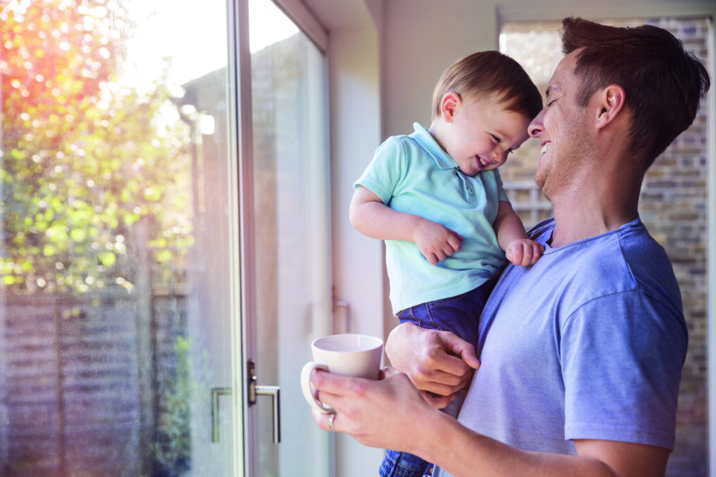 Father holds toddler son while drinking coffee at home, by the window